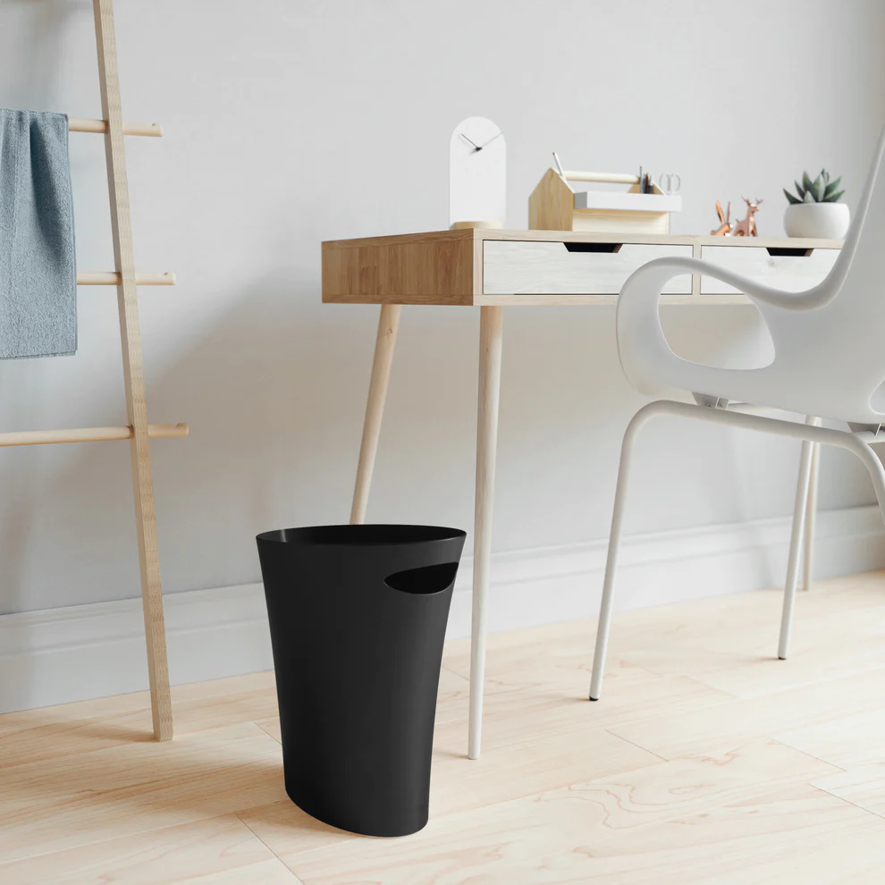 Black skinny trash can by Umbra, shown next to a wooden desk